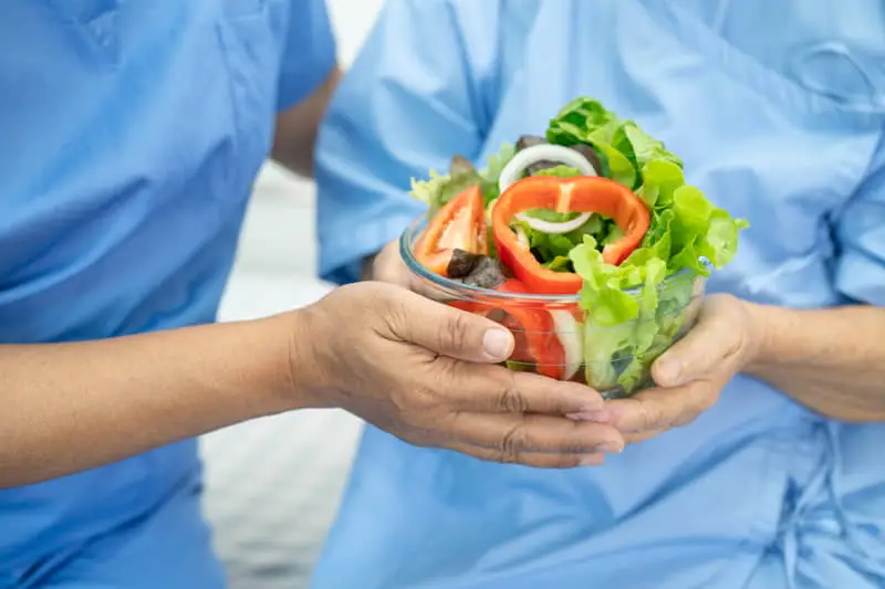 Dos personas con uniforme médico azul sosteniendo un tazón transparente con ensalada fresca de lechuga, pimiento rojo y tomate.