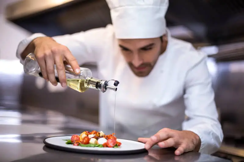 Chef vertiendo aceite sobre un plato con tomates cherry y mozzarella en una cocina profesional.